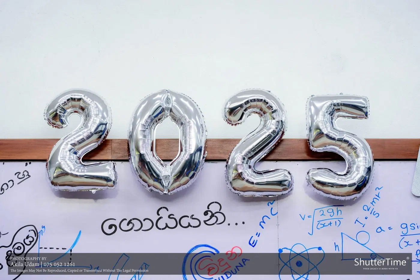 රිදී පැහැති 2025 බැලූන් අකුරු, පසුබිමේ ලියූ වයිට් බෝඩ් එකක්