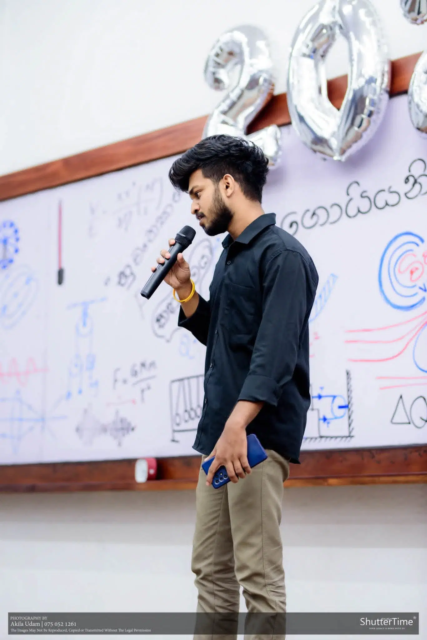 සිසුවෙක් වයිට් බෝඩ් එකක් ඉදිරිපිට මයික්‍රොෆෝනයක් අතැතිව