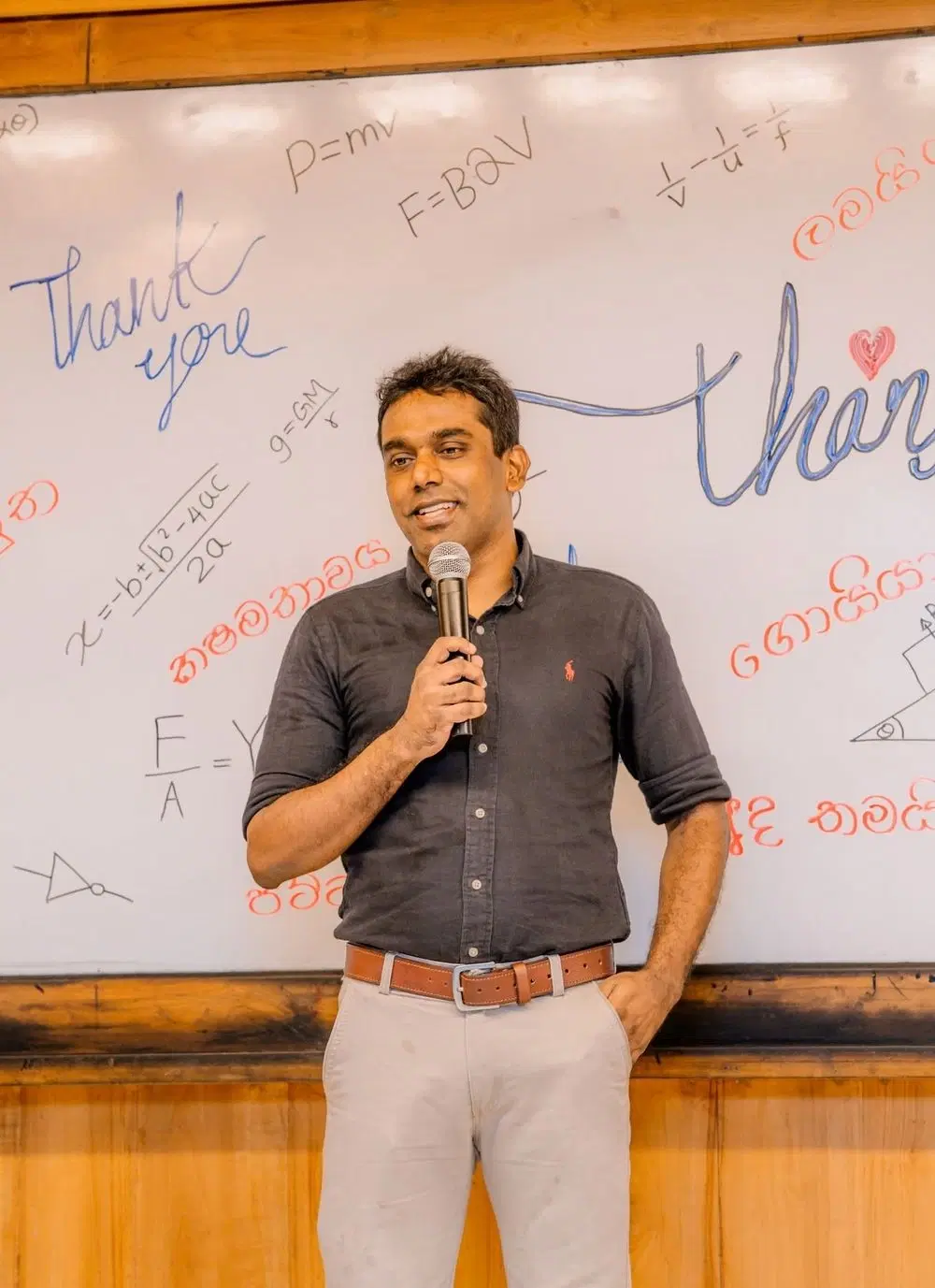 සුජිත් ලියනගේ, භෞතික විද්‍යා සමීකරණ සහ සිසුන් අතින් ලියූ ස්තුති පණිවිඩ සහිත වයිට් බෝඩ් එක ඉදිරිපිට, මයික්‍රෆෝනයක් අතැතිව